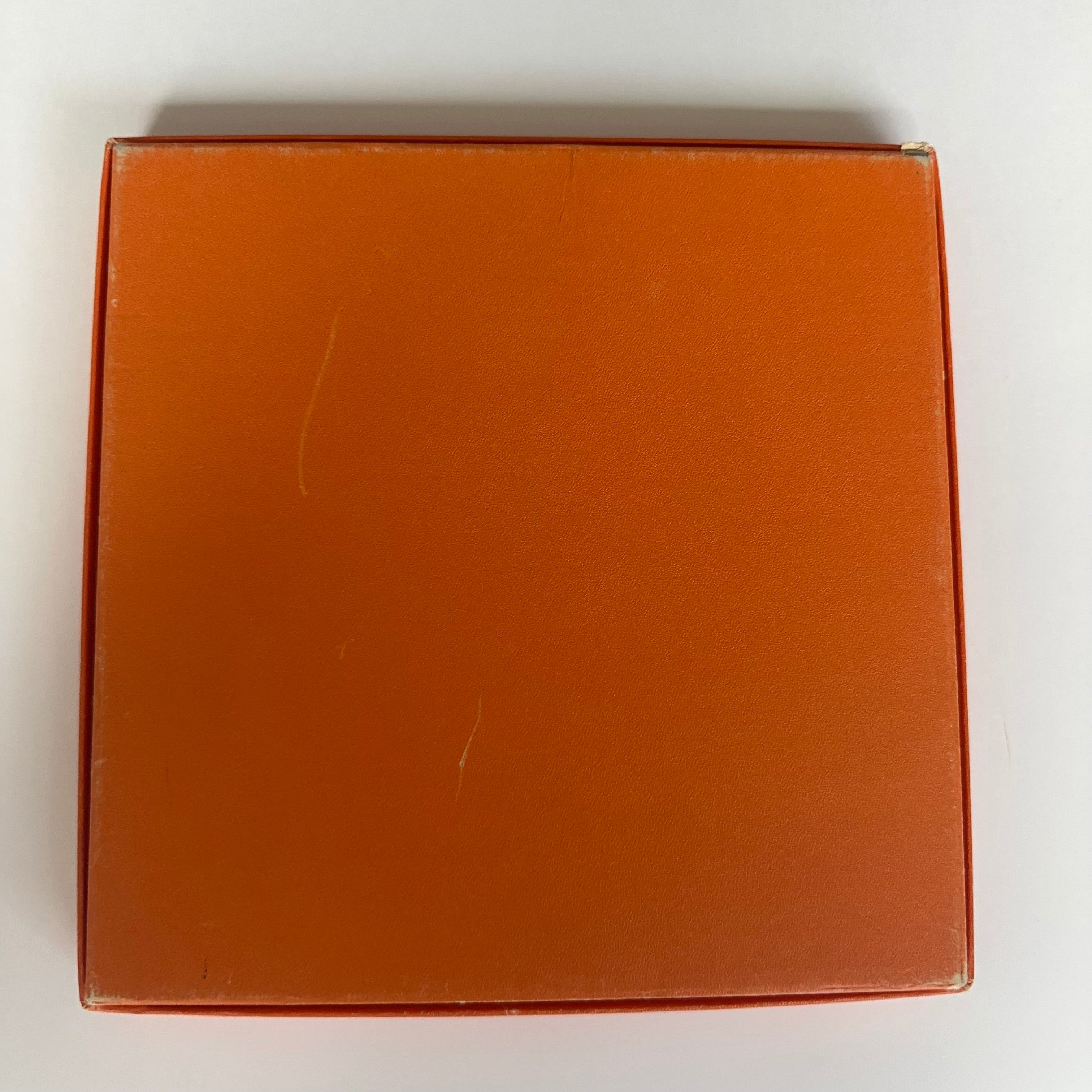 Vue de détail montrant de petits défauts sur le fondde la boîte orange Hermès fournie avec le foulard (mentionnés dans la description).