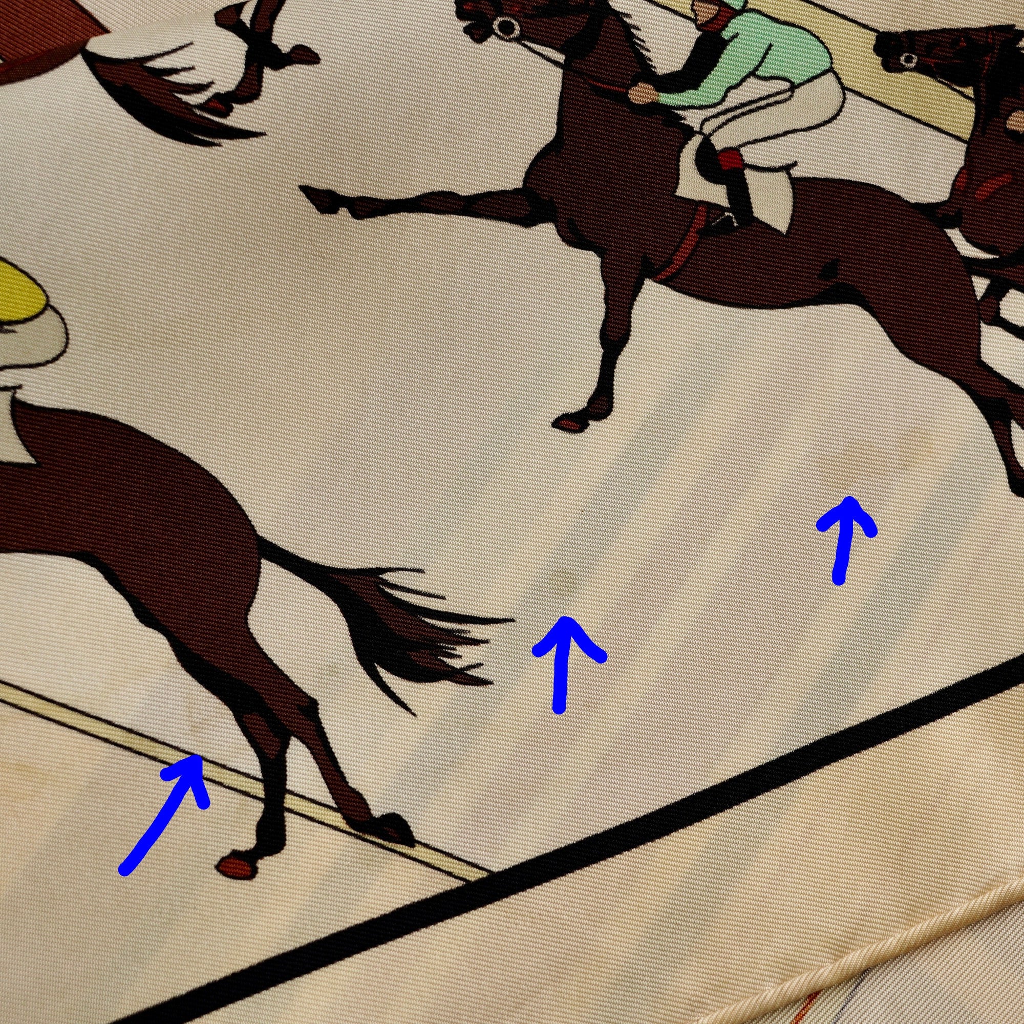Zoom sur de petites taches sur le fond du foulard Hermès "Les Courses", pointée par trois flèches bleues.